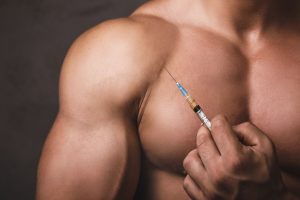 A close-up of a muscular man holding a syringe filled with steroid oil near his chest against a dark background.