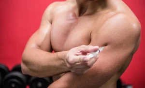 Bodybuilder self-injecting testosterone into the deltoid muscle against a red background with gym equipment.