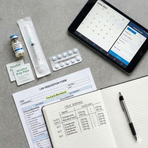 A flat-lay photograph of a steroid cycle planning setup featuring a testosterone vial, syringe, alcohol prep pads, PCT pills, a lab requisition form, an open notebook with a handwritten cycle schedule, and a tablet displaying a calendar app.